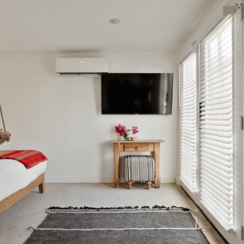 Relax in this bright, airy bedroom featuring a cozy bed with vibrant textiles, a rustic wooden table , and plenty of natural light streaming through elegant blinds, your perfect retreat awaits.